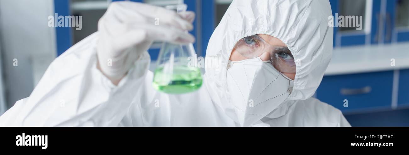 Scientist in hazmat suit and protective mask holding flask with liquid ...