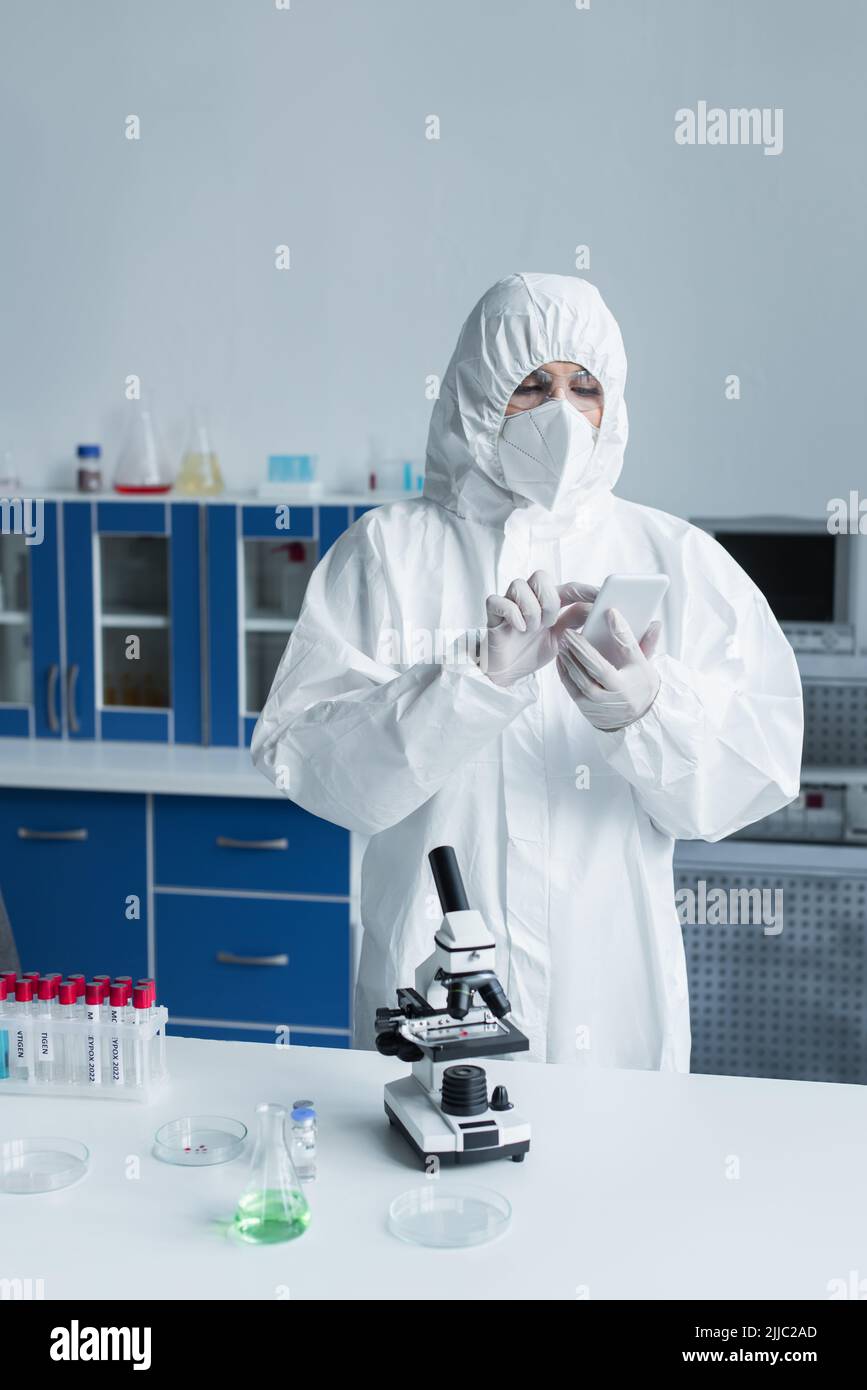 Scientist in hazmat suit using smartphone near microscope and test tubes in lab Stock Photo