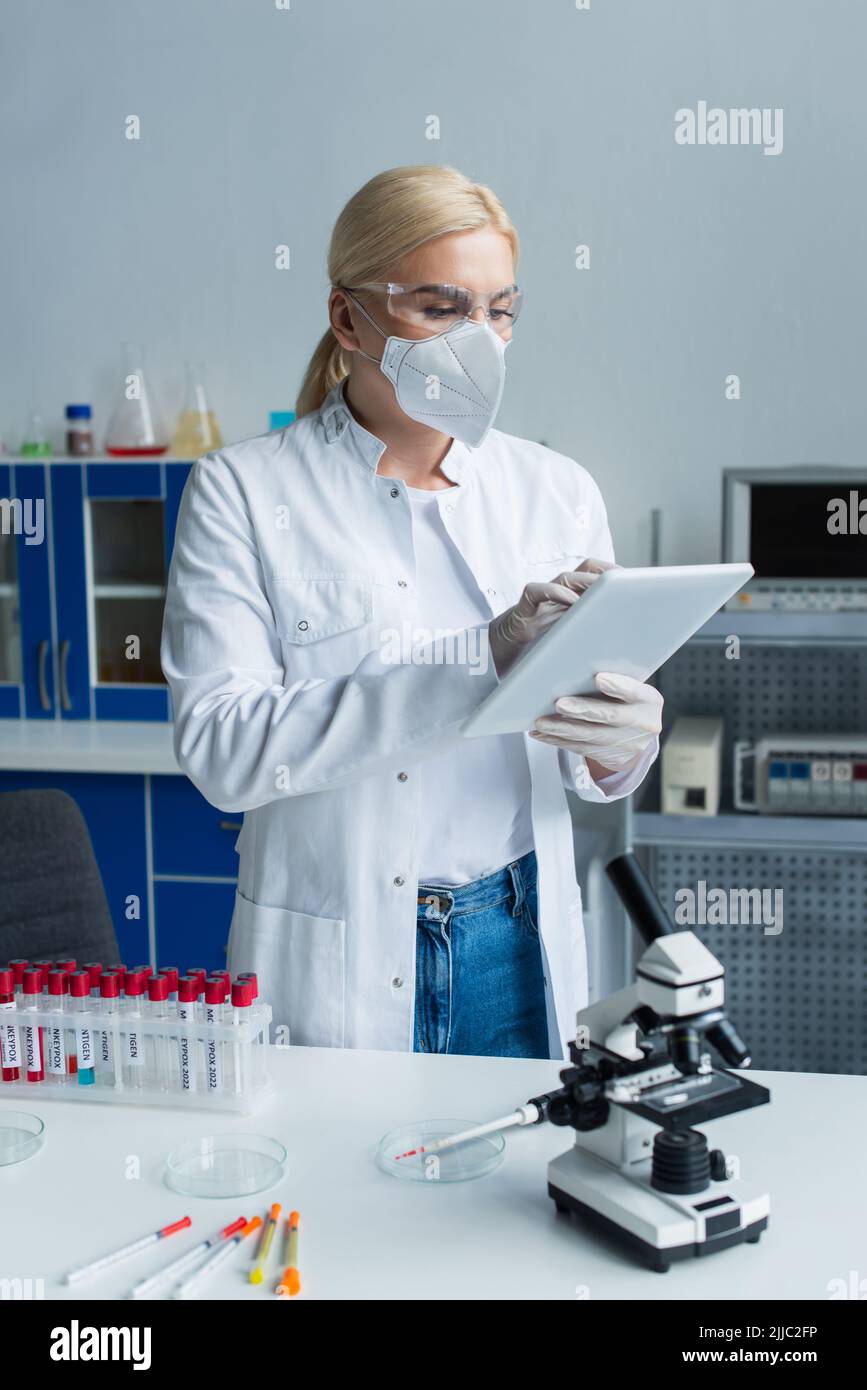 Scientist in protective mask using digital tablet near test tubes and microscope in lab Stock Photo