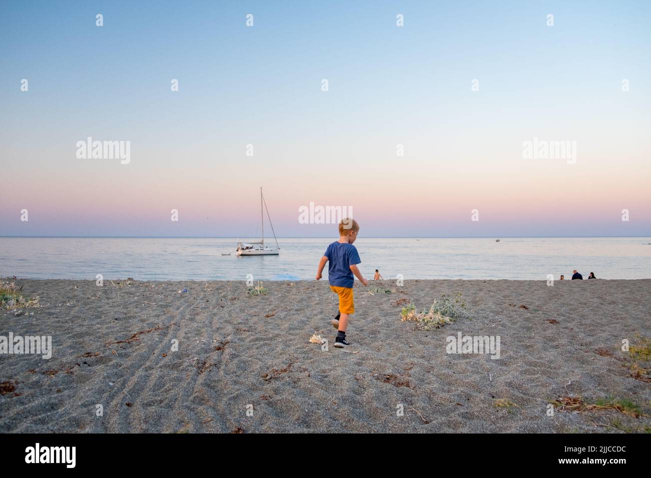 Cute little boy staring a sailboat anchored near the shore of Agiokampos beach at sunset time , Greece Stock Photo