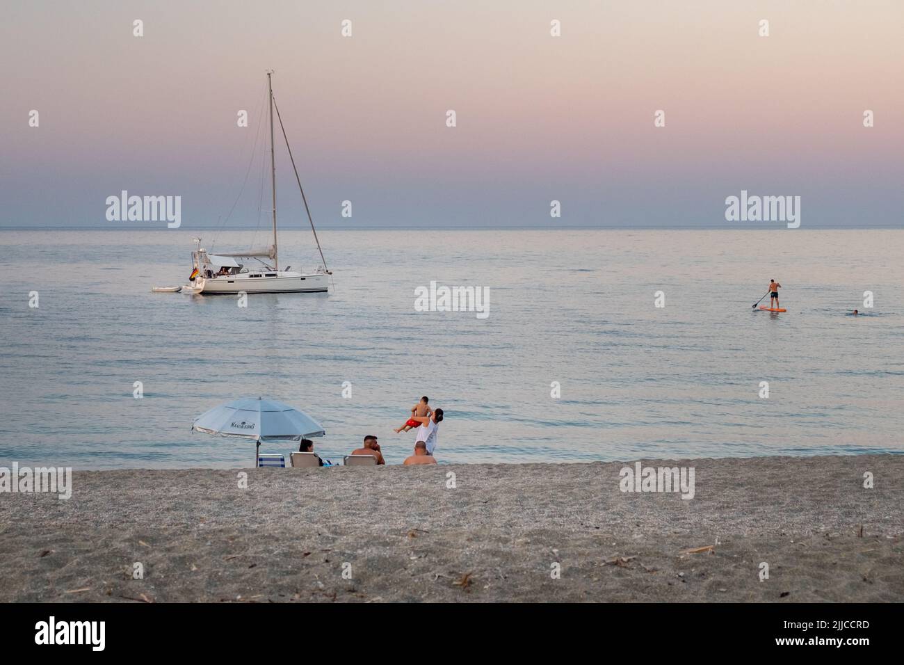 People at the beach sitting, staring and stant up boarding at sunset time with a sailboat anchored near to the shore of Agiokampos beach , Greece Stock Photo