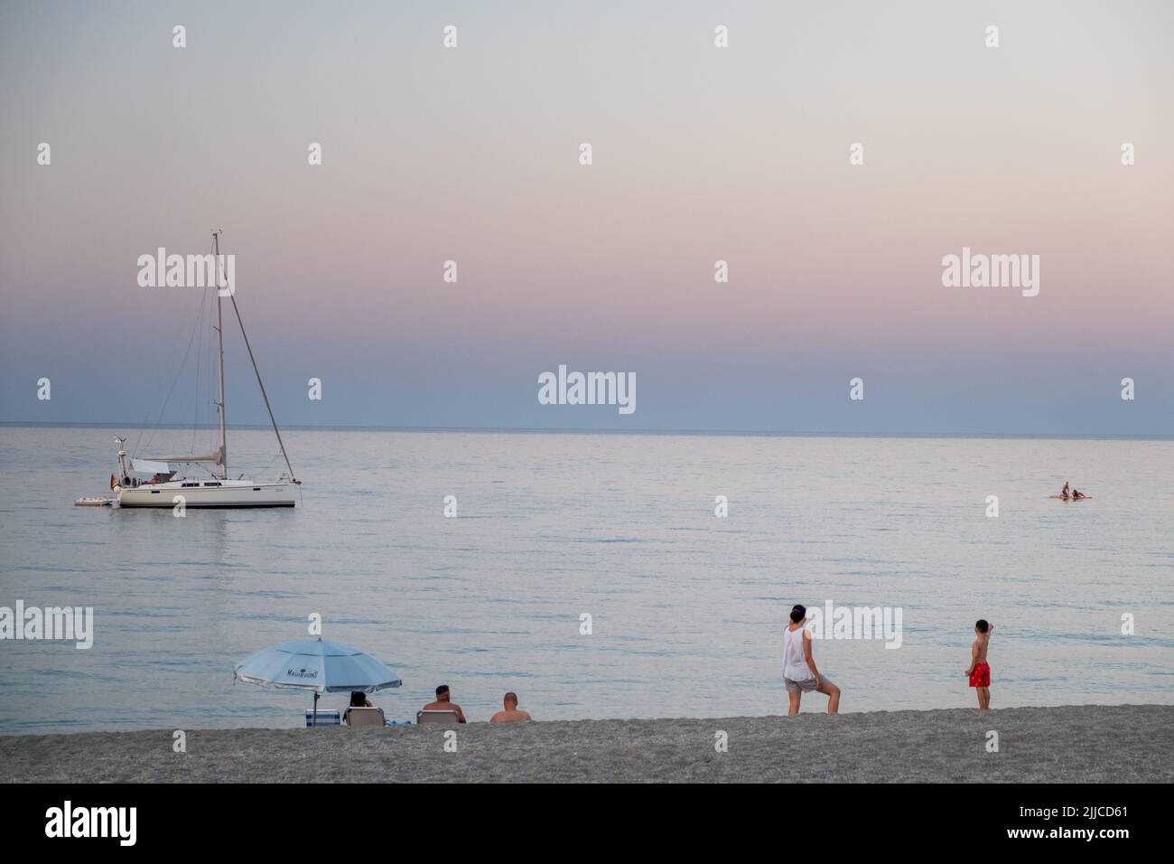 People at the beach sitting staring the sunset and the sailboat at Agiokampos beach , Greece Stock Photo