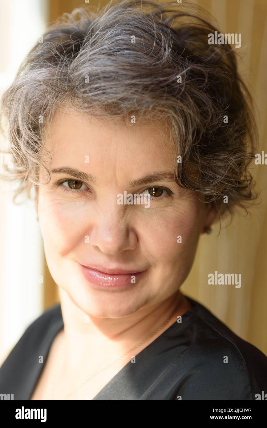 Head and shoulders portrait of cheerful and slyly smiling gray haired woman looking at camera. Shot indoors with natural side lighting from window. Stock Photo