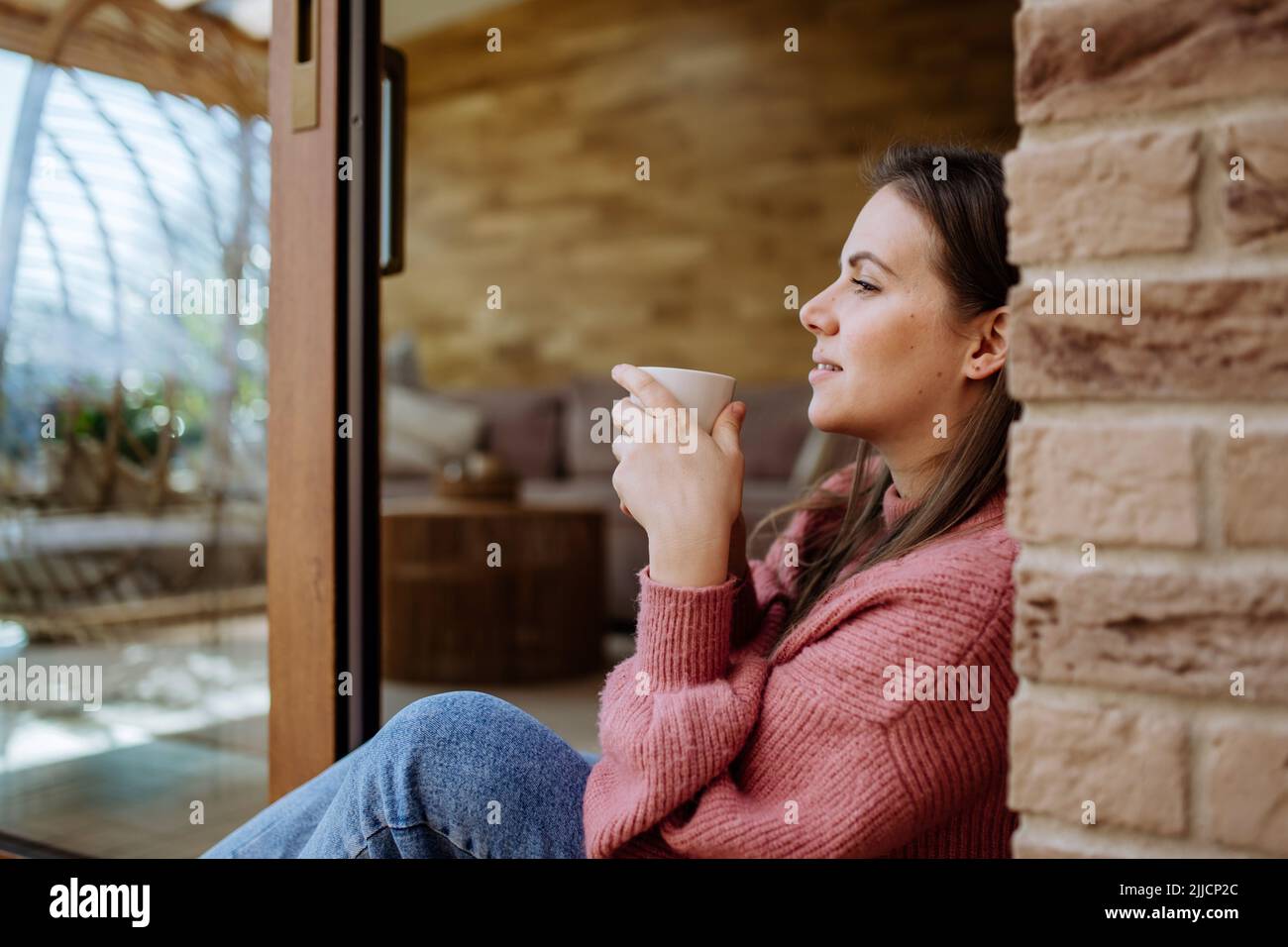 Smiling young woman with dreamy face drinking morning coffee and sitting at her home patio Stock Photo