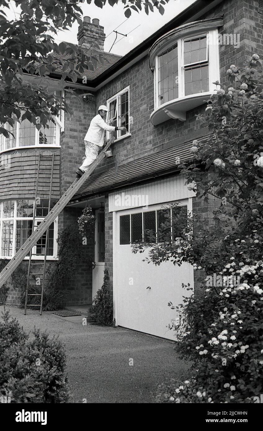 1970s, at the front of a large suburban house. a man in a white decorator's suit and with cap, standing on a ladder painting the wooden frame of a first floor window, England, UK. Stock Photo