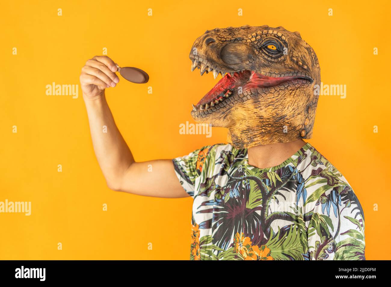 Man in dinosaur animal head mask eating chocolate ice cream isolated on yellow background with copy space Stock Photo