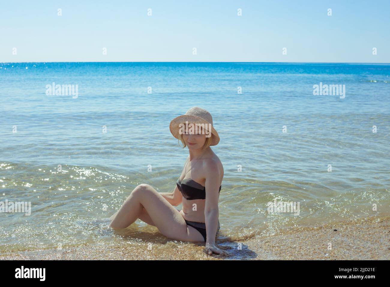 An adult slender woman with fair skin in a swimsuit sits in sea water. Summer vacation, rest and relaxation on the sea coast. Stock Photo