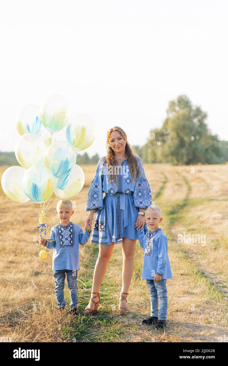 Happy mother with two twins sons walk on meadow with air balloons. They are dressed in Ukrainian national embroidered shirts. Stock Photo