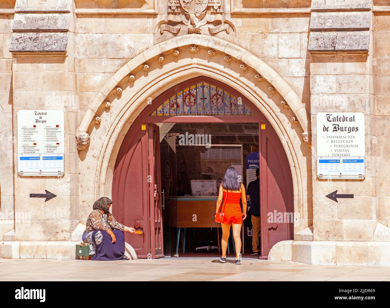 Woman in a red dress at the  entrance to the cathedral of St Mary the virgin in the Spanish city of Burgos Spain with a beggar asking for money Stock Photo