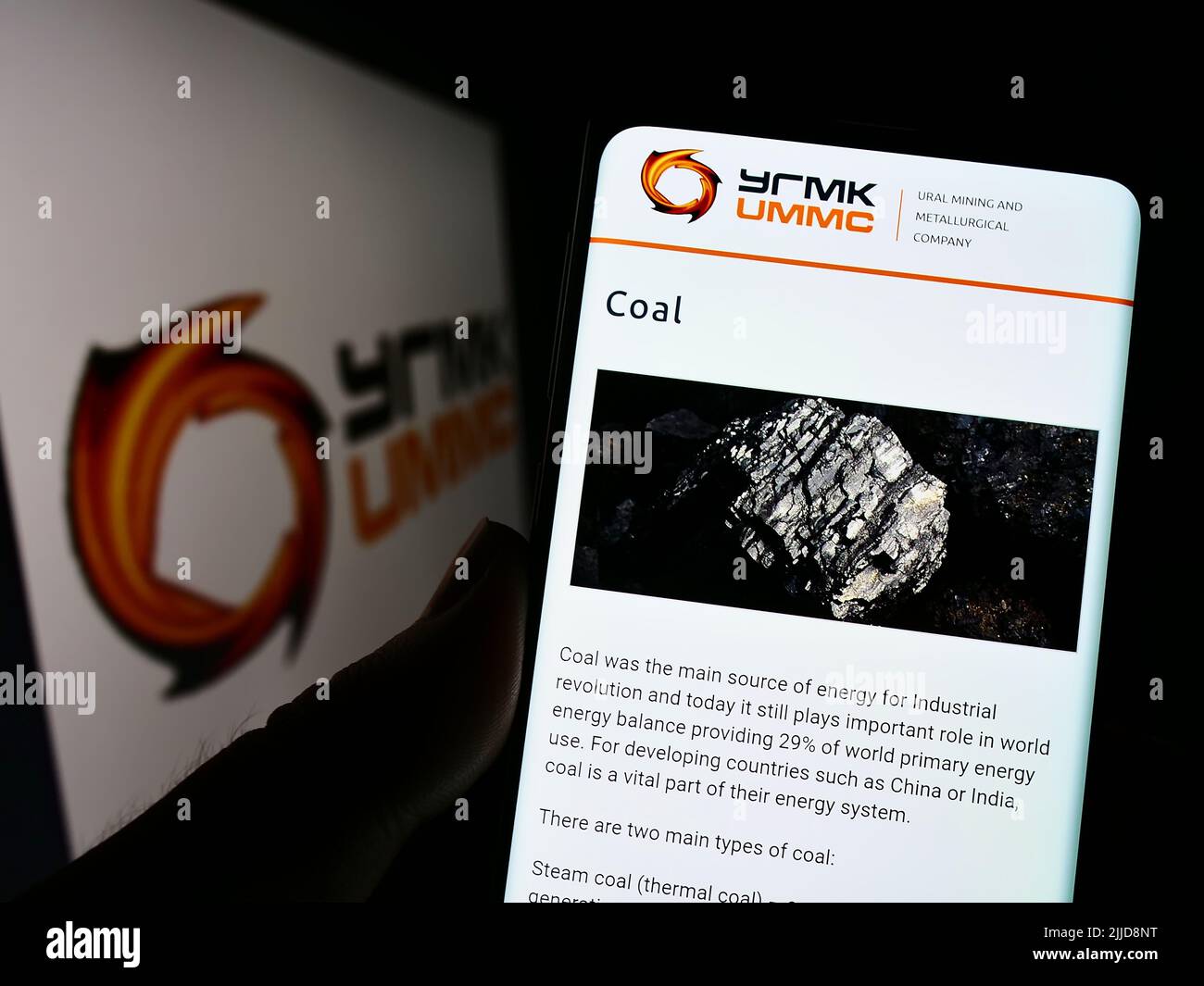 Person holding cellphone with webpage of Ural Mining and Metallurgical Company (UMMC) on screen with logo. Focus on center of phone display. Stock Photo