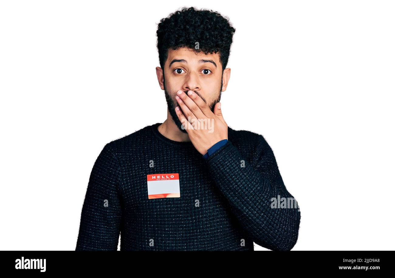 Young arab man with beard wearing hello my name is sticker identification covering mouth with hand, shocked and afraid for mistake. surprised expressi Stock Photo