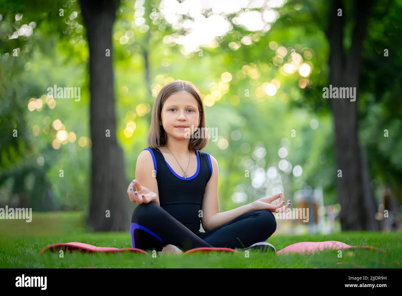 a little girl sitting in a park meditates in a folded lotus position ...