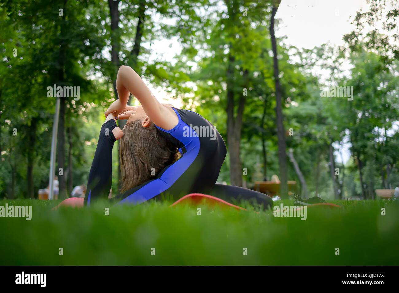 a little girl sitting on the lawn in the park performs backbends she does yoga elements to stretch her back Stock Photo