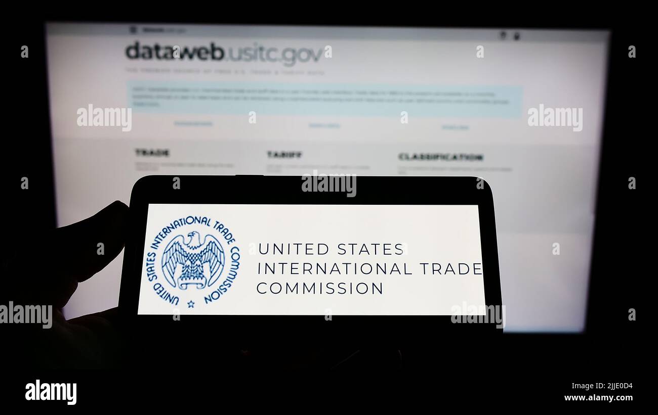 Person holding mobile phone with logo of US International Trade Commission (USITC) on screen in front of web page. Focus on phone display. Stock Photo