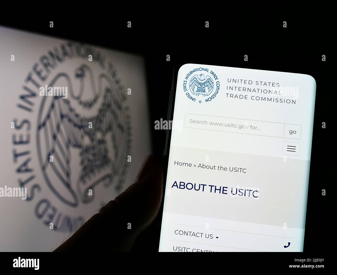 Person holding cellphone with website of US International Trade Commission (USITC) on screen in front of logo. Focus on center of phone display. Stock Photo