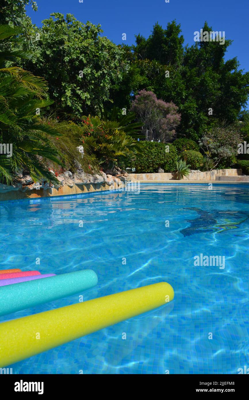 Bright and colourful swim noodles floating on bright blue swimming pool in Mediterranean garden in summer Stock Photo