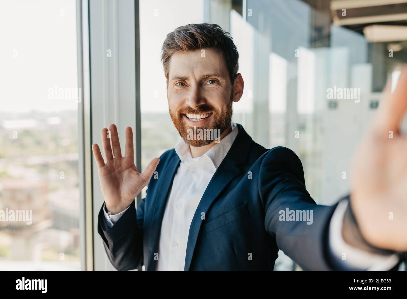 Happy successful businessman with beard in suit waving his hand at smartphone camera, having remotely meeting Stock Photo