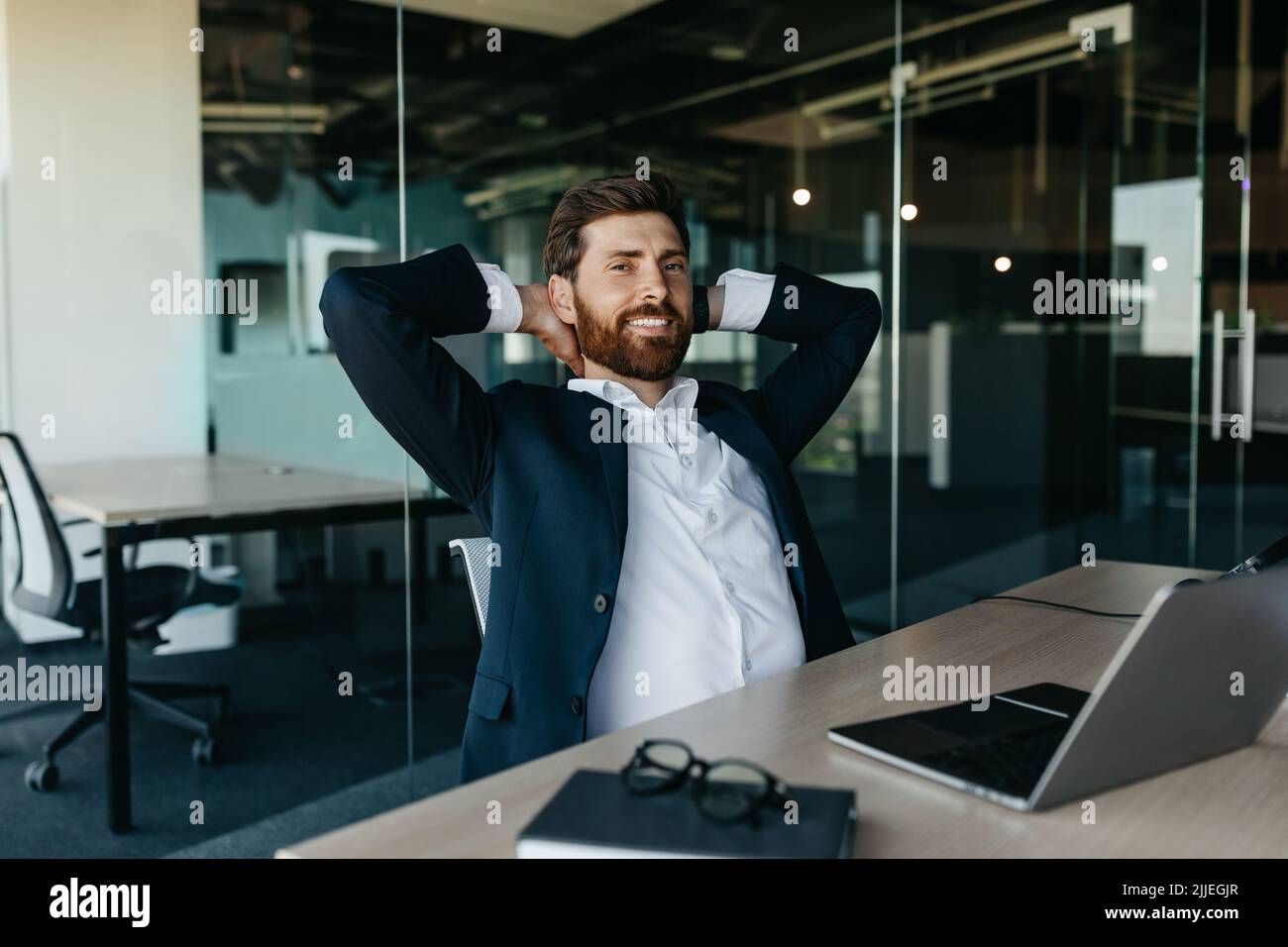 Relaxed happy businessman sitting at workplace in office, leaning back on chair and smiling, free space Stock Photo