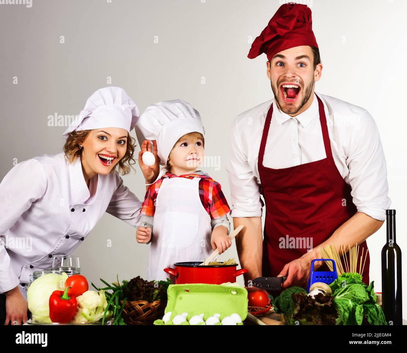 Homemade food. Family preparing dinner in kitchen. Little son and parents in uniform and chef hat. Stock Photo