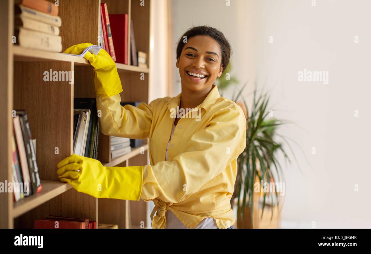 African Woman Dusting And Cleaning Bookshelf With Rag At Home Stock Photo