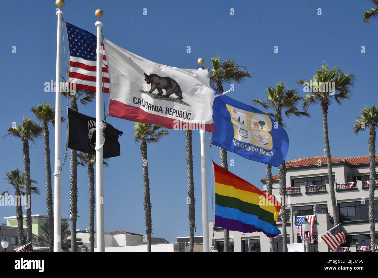 Several flags flying at Huntington Beach in front of palm trees against a cloudless blue sky, including Stars & Stripes and California Republic flags. Stock Photo