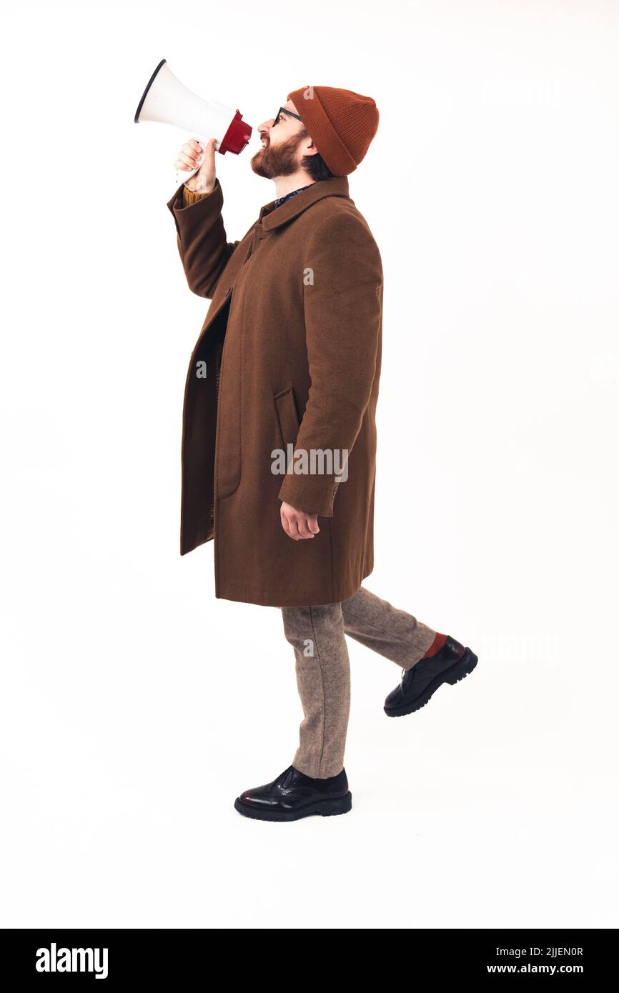 Man in his 30s using megaphone to yell - dressed in a camel coat and stylish boots - profile shot on white background - isolated. High quality photo Stock Photo