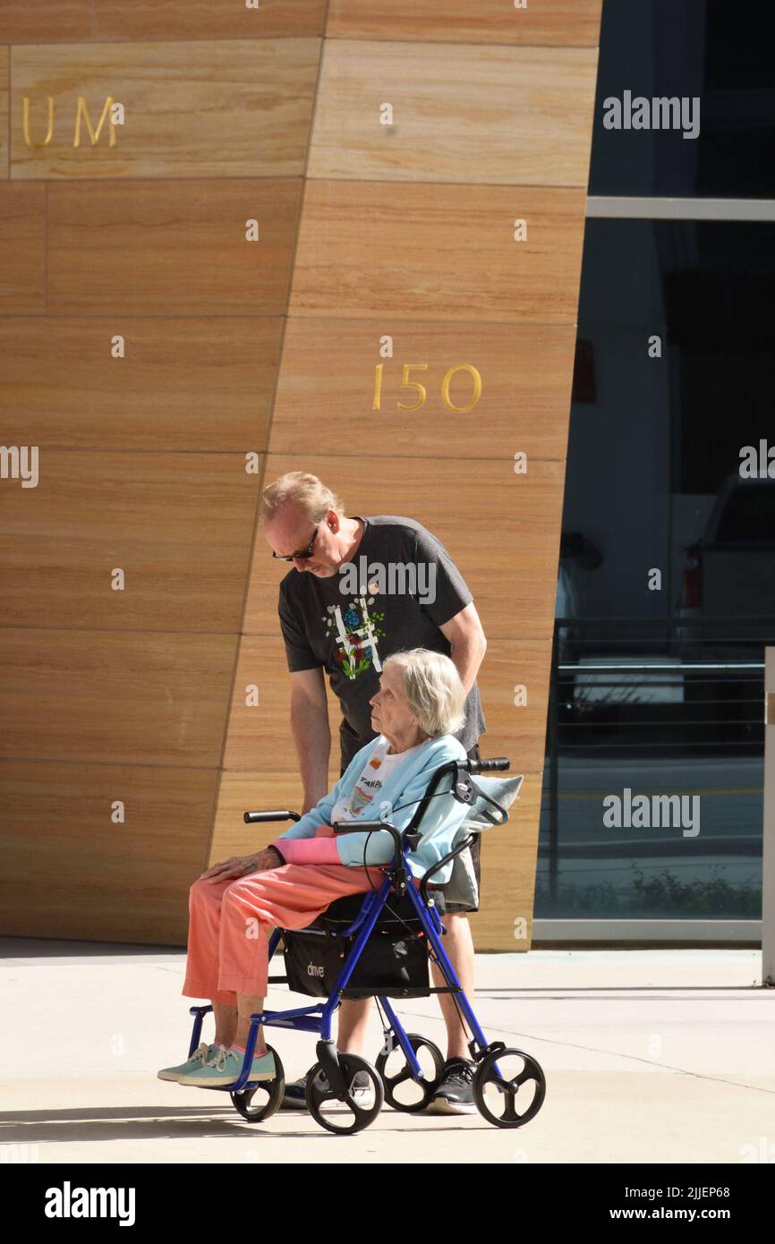 A Caucasian senior man assisting an elderly woman who is sitting on the rollator wheelchair in front of the building on a sunny day. Stock Photo
