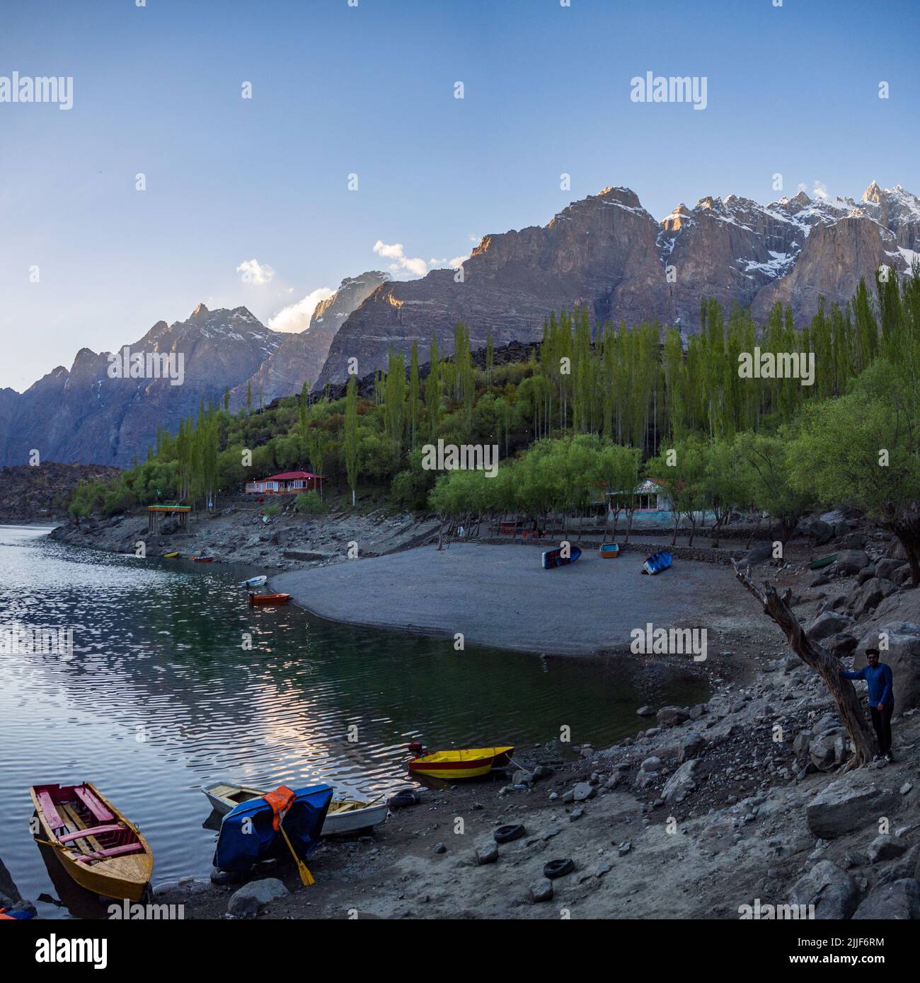 The Upper Kachura lake with a forest and boats, a male on the coast surrounded by rocky mountains in Skardu, Pakistan Stock Photo
