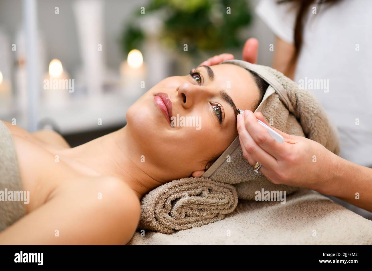 Crop anonymous beautician tweezing eyebrows of content female client lying on couch during beauty routine in spa salon against blurred background Stock Photo