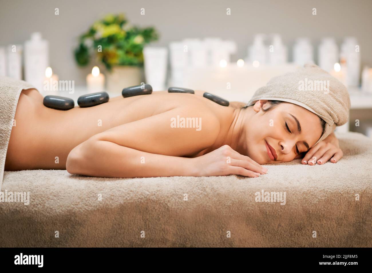 Pleased female client with hot stones on back lying on couch with towel during massage session in modern light spa Stock Photo