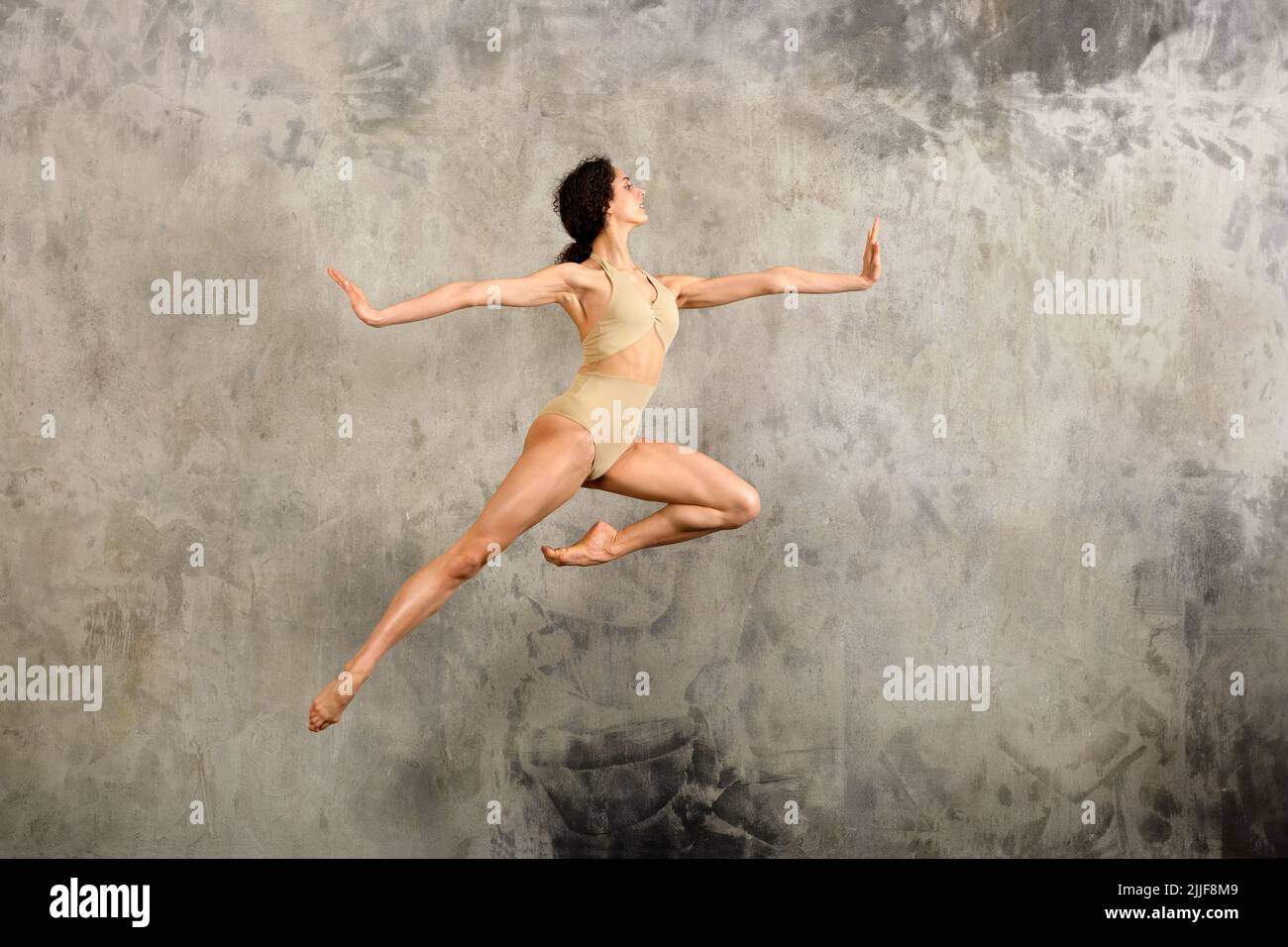 Full body side view of female ballet dancer in bodysuit leaping up with spread arms against gray wall in studio Stock Photo