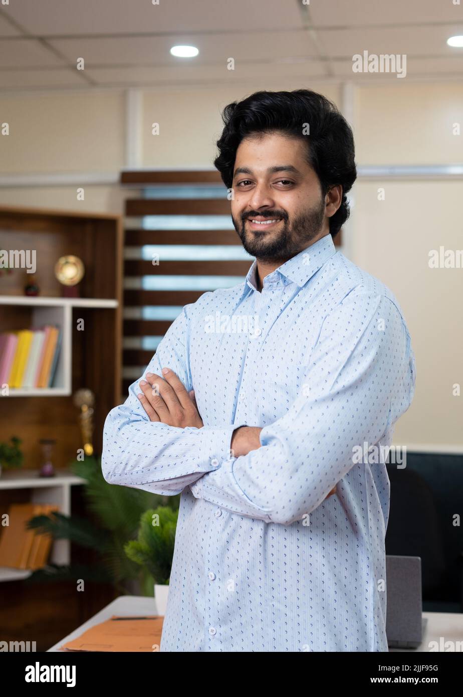 Vertical shot of smiling young businessman at office confidently standing with crossed by arms looking at camera - concept successful entrepreneur Stock Photo
