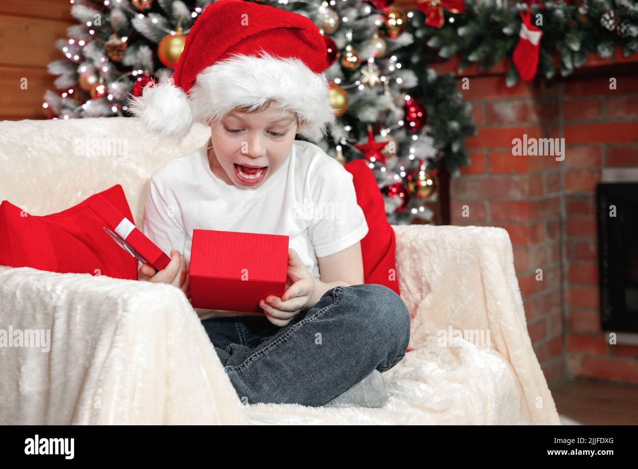 Merry Christmas kids. Emotions of a child. Joyful surprised kid boy in Santa's hat opened a Christmas box with a gift from Santa Claus near the Christ Stock Photo