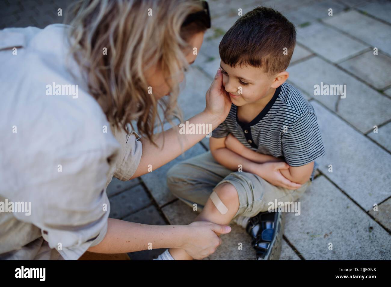 Young mother comforting her little son and giving him bandage after getting hurt when fall down in street. Stock Photo