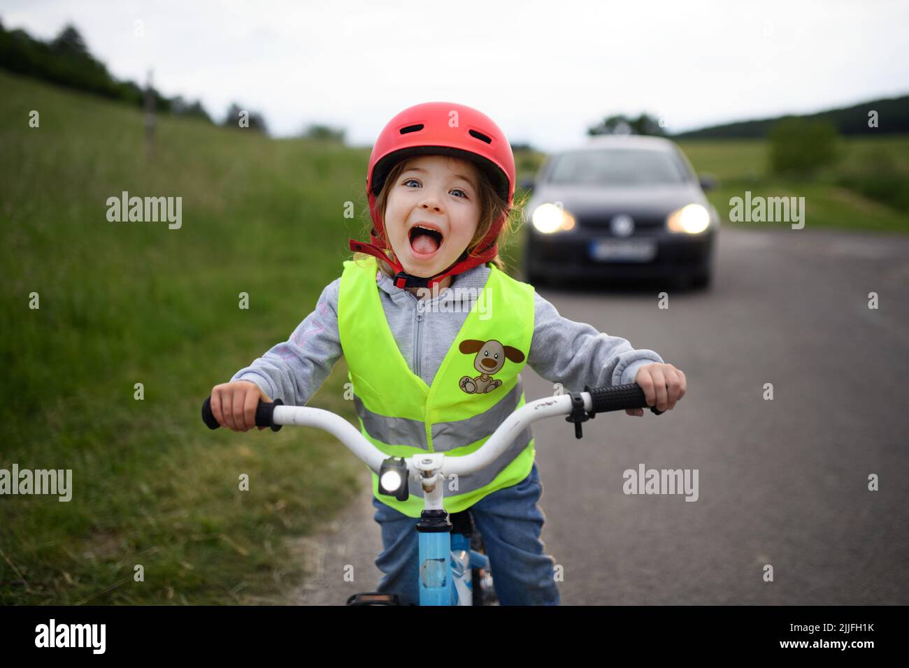 Portrait of excited little girl in reflective vest riding bike on road with car behind her, road safety education concept. Stock Photo