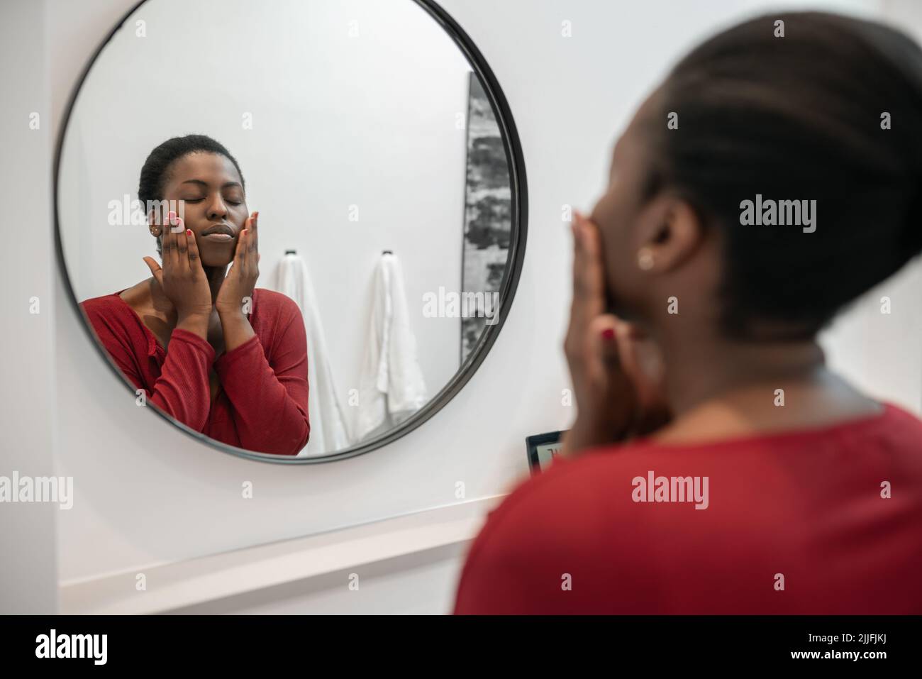 Bathroom mirror reflection of Beautiful Black African woman touching her face, applying moisturizer, eyes closed . Part of her morning self care routi Stock Photo