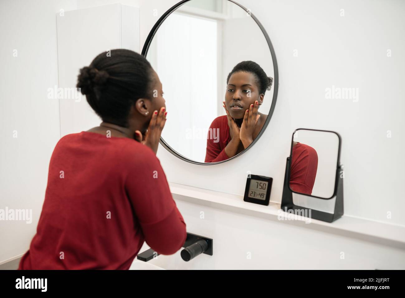 Beautiful Black African woman looking at her face in the bathroom mirror hands on cheeks. Part of her morning self care routine Stock Photo