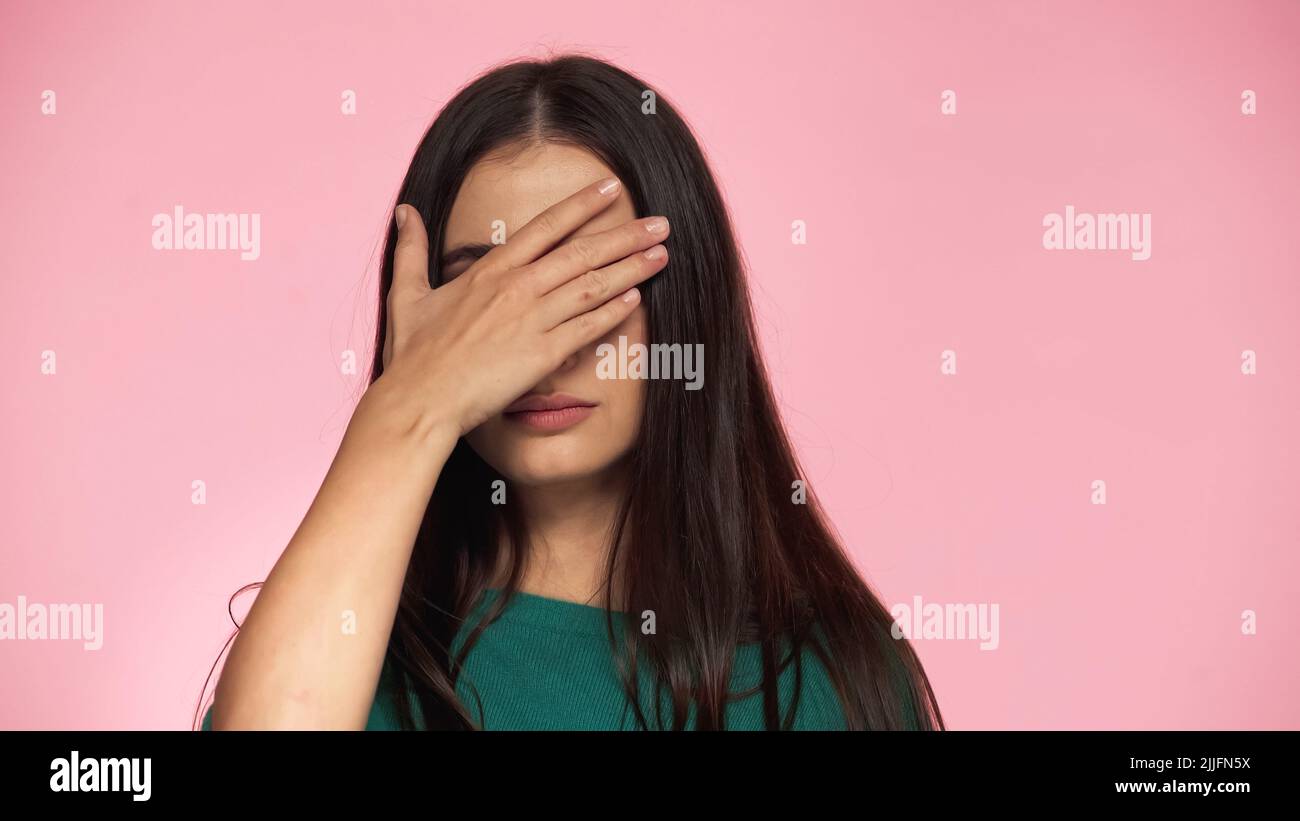 brunette young woman in green blouse covering eyes with hand isolated on pink Stock Photo