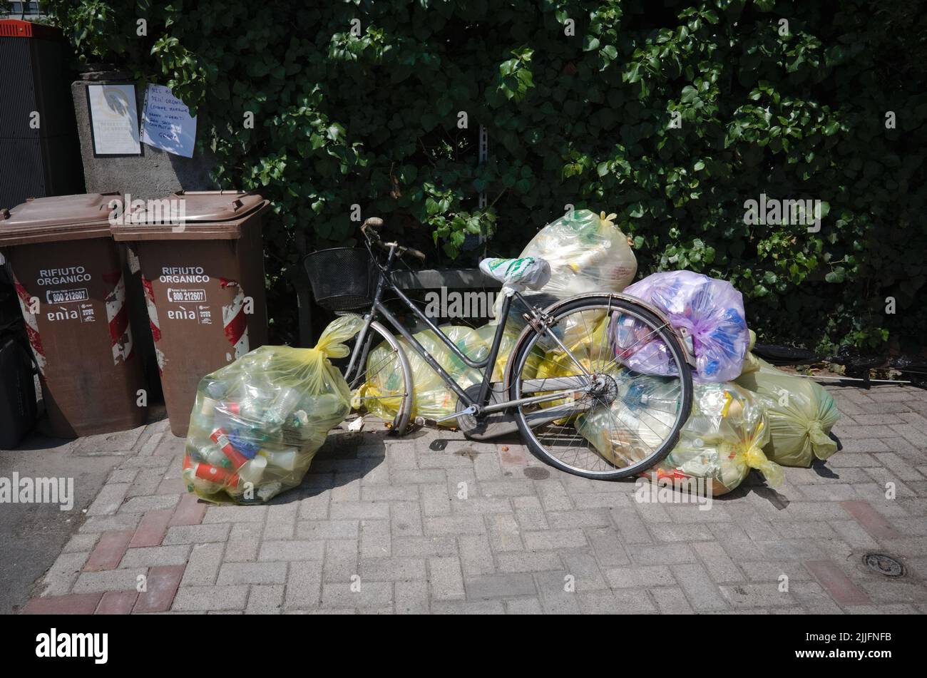 Parma, Italy - July, 2022: Old bicycle with basket on handlebar and broken wheel among garbage bags with plastic near brown trash cans. Discarded bike Stock Photo