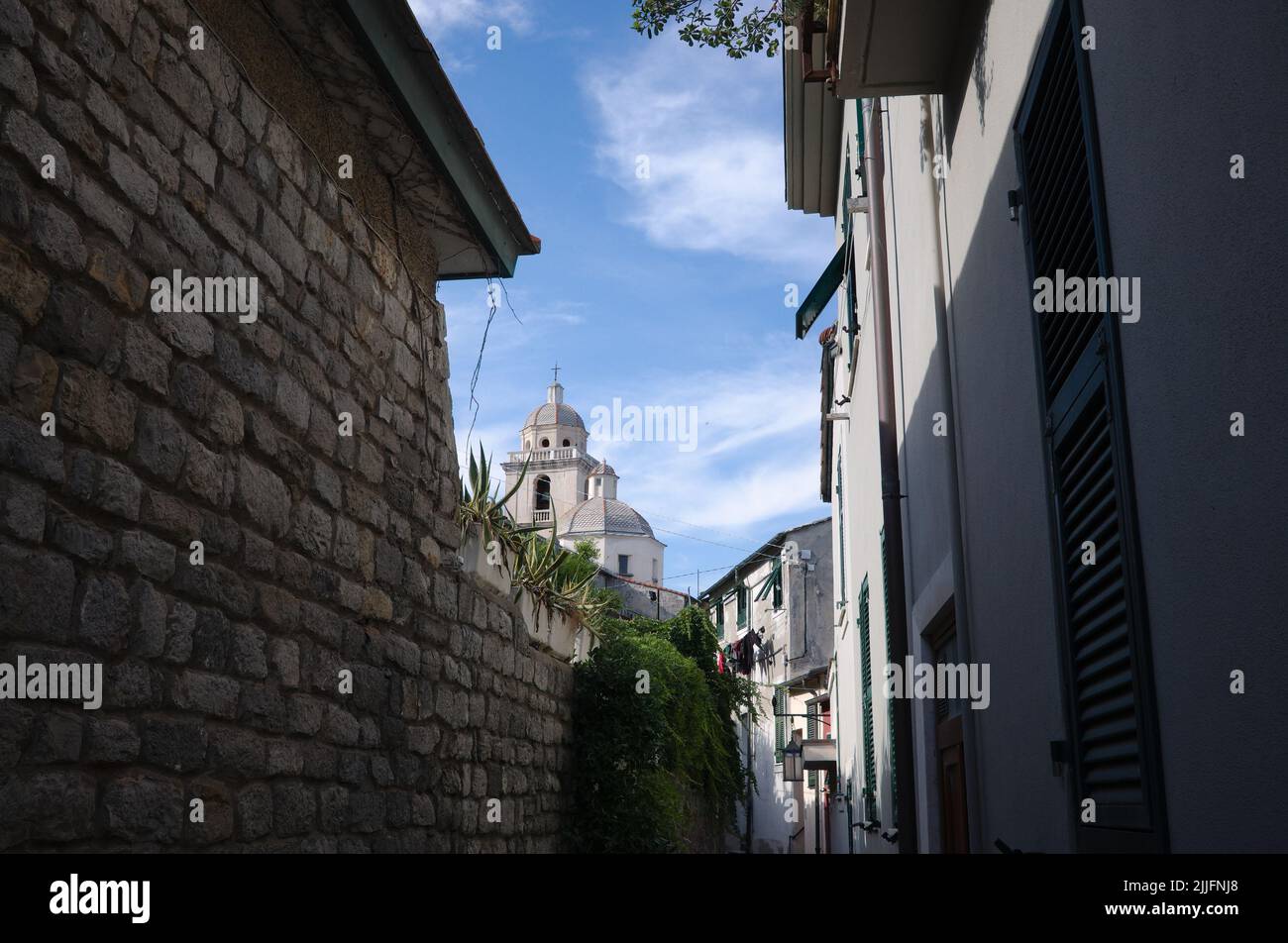 Narrow street with view to San Lorenzo church in Porto Venere, Liguria, Italy. Passage between stone wall and Italian houses with shutters on windows. Stock Photo