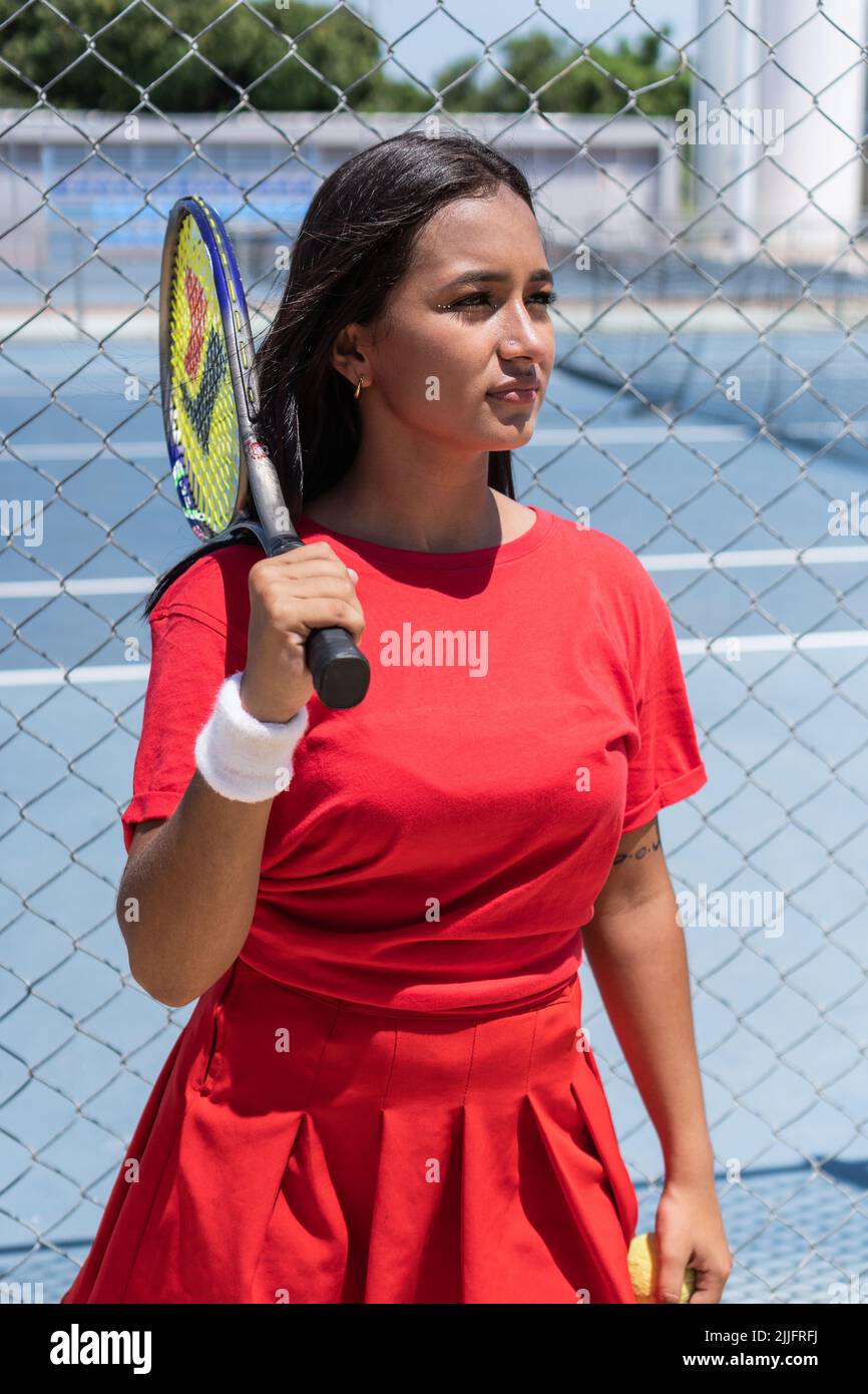 Brunette Woman with racket and ball standing near tennis court fence during break in training. Stock Photo