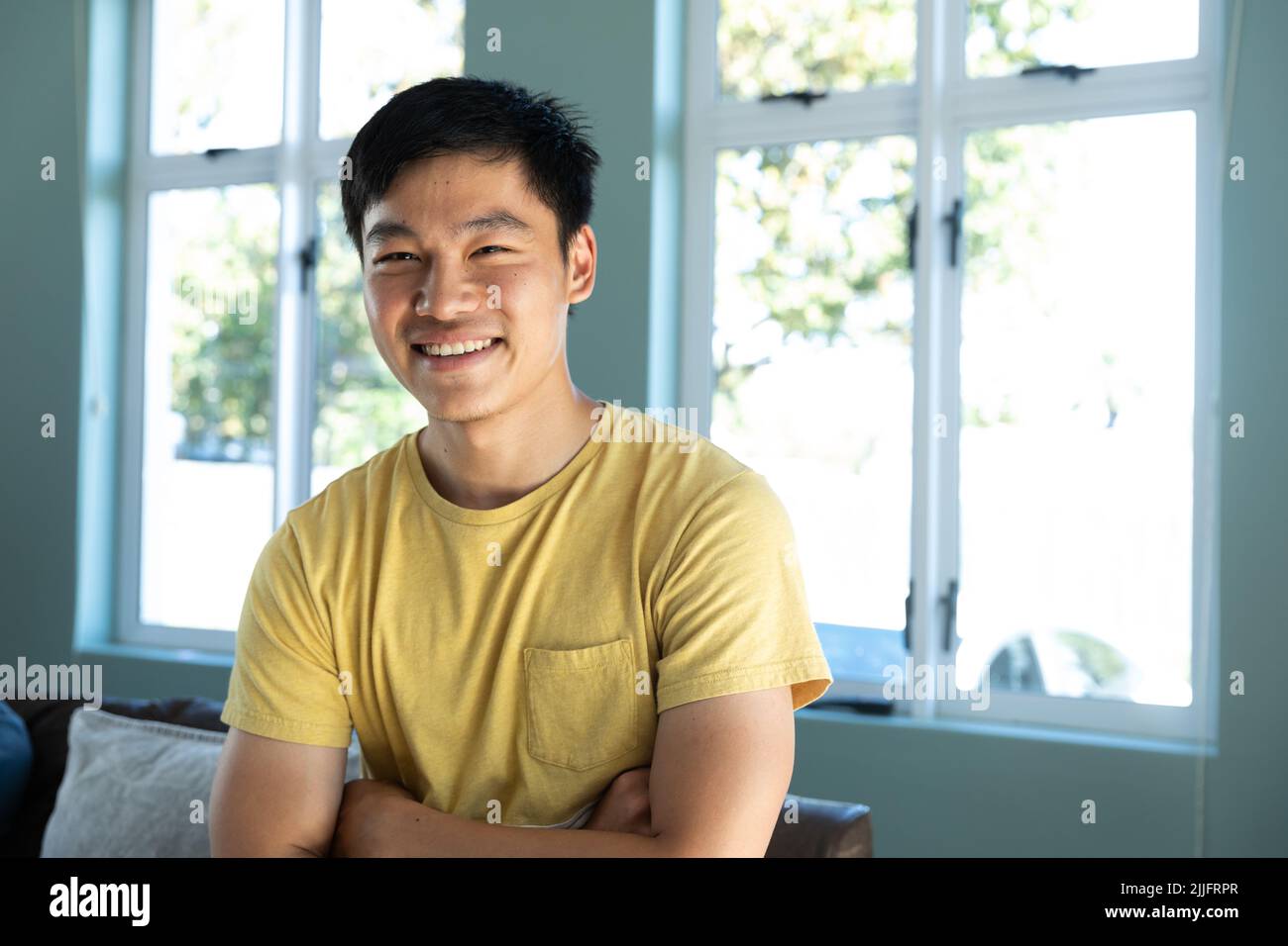 Portrait of smiling chinese young man with arms crossed standing against windows at home Stock Photo