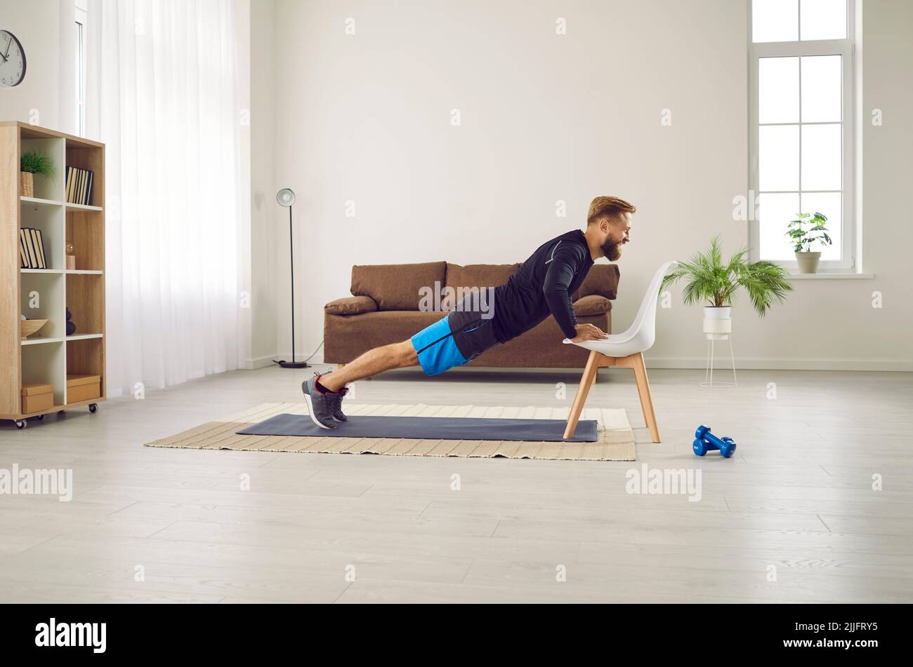 Athletic man is doing push up exercise with chair at home in his living room with modern interior. Stock Photo