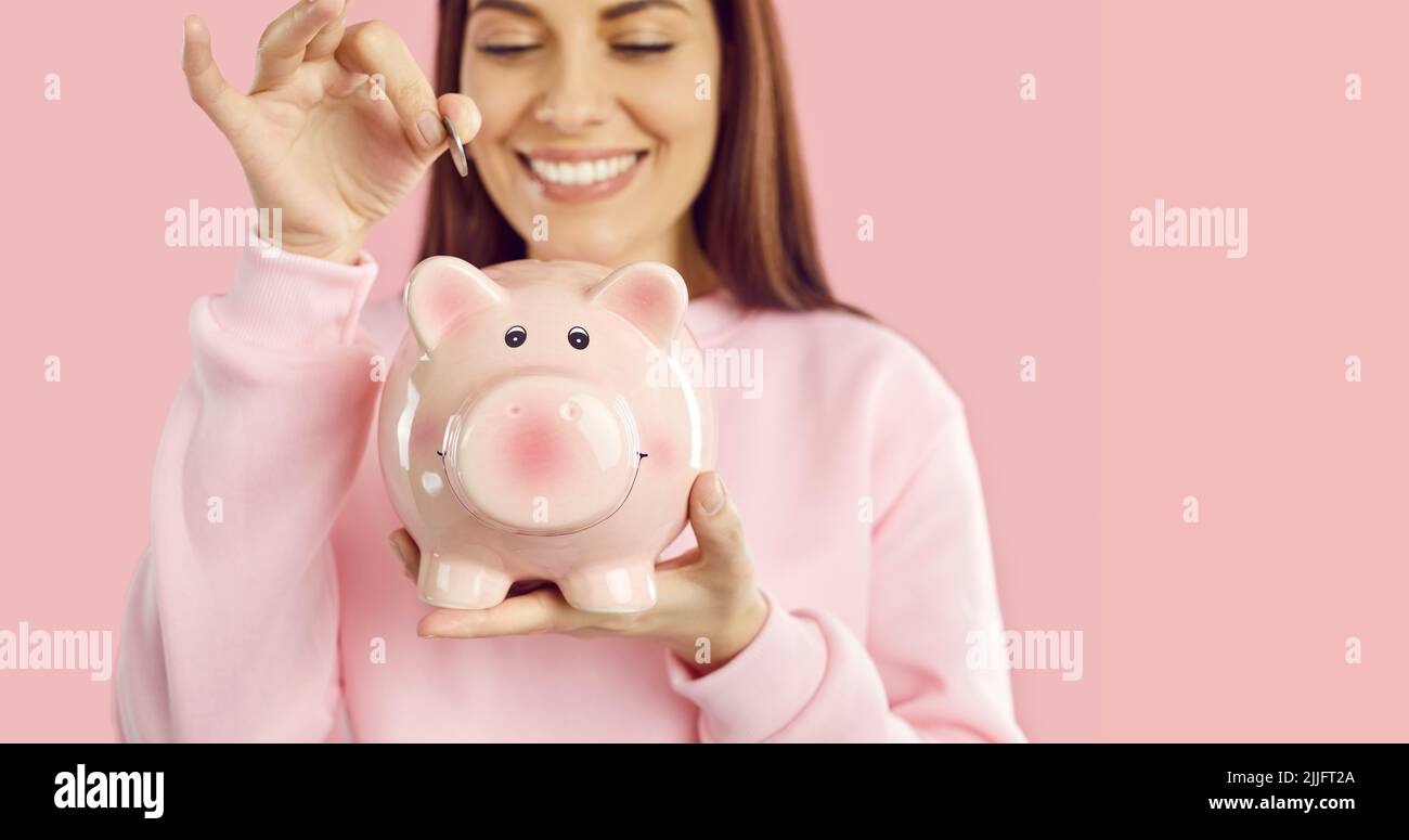 Happy young woman who is saving up some money puts a coin in her pink ...