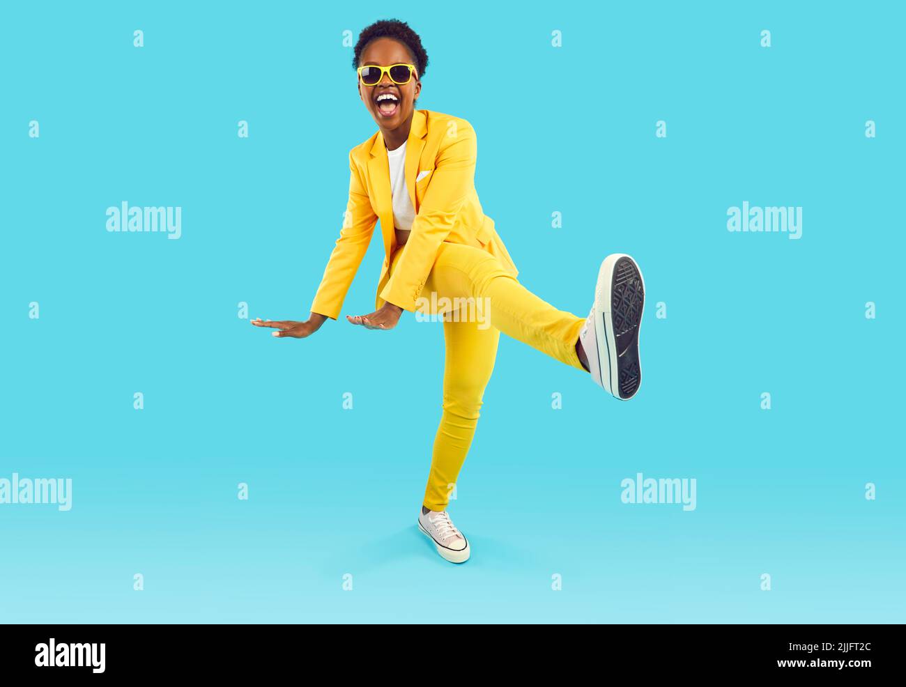 Happy African American woman in yellow suit and sunglasses dancing isolated on blue background Stock Photo