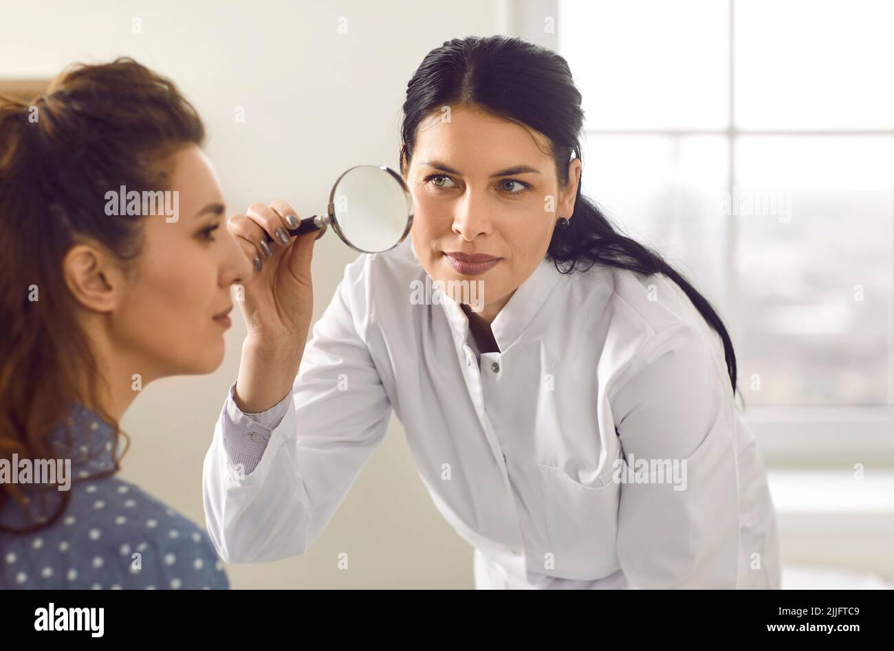 Dermatologist with magnifying glass looks at mole or pigmentation on woman's face Stock Photo