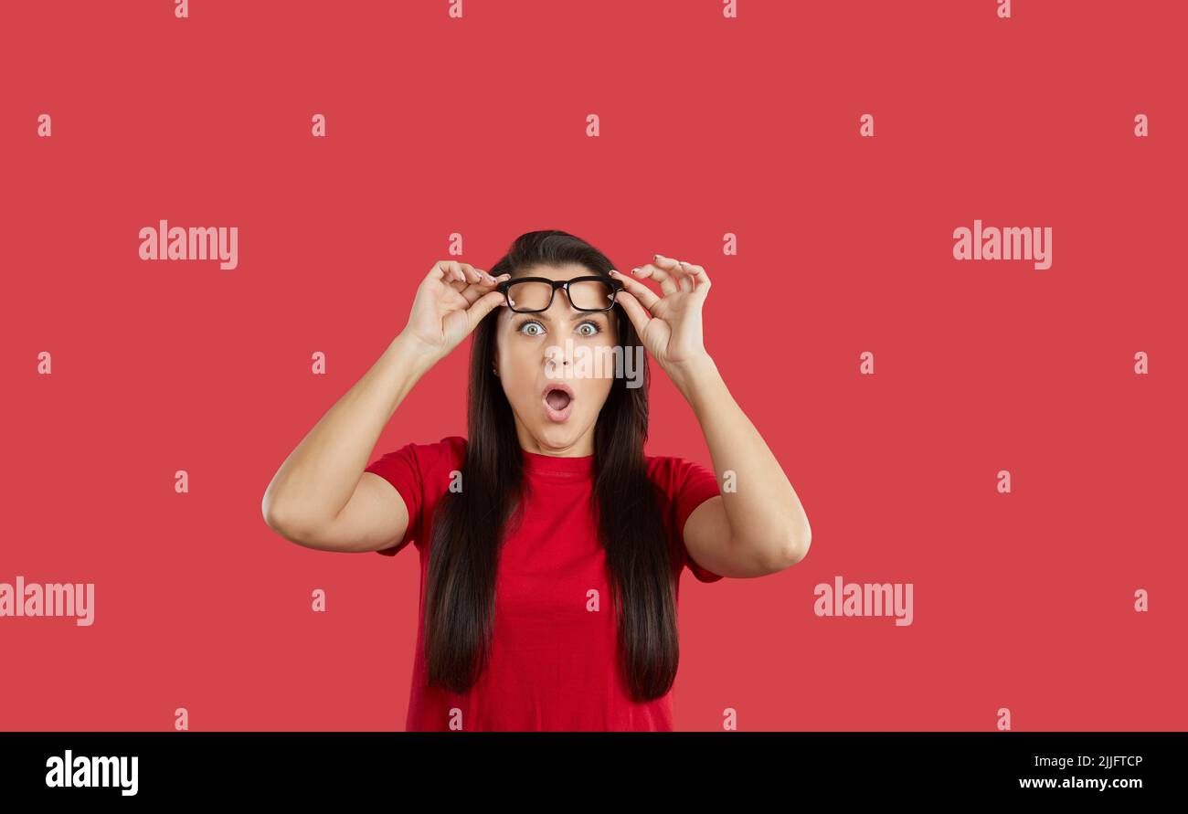 Surprised and shocked caucasian girl with eyeglasses, with eyes widened and mouth opened, holding glasses with hands on forehead, over red background Stock Photo