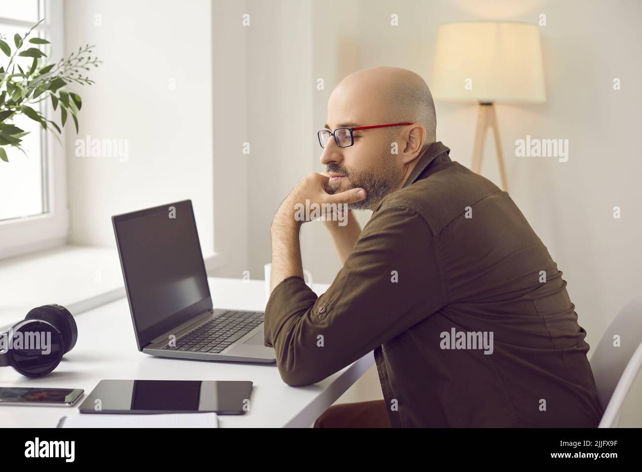 Pensive man sitting at his working desk with laptop, tablet, mobile phone and headphones Stock Photo