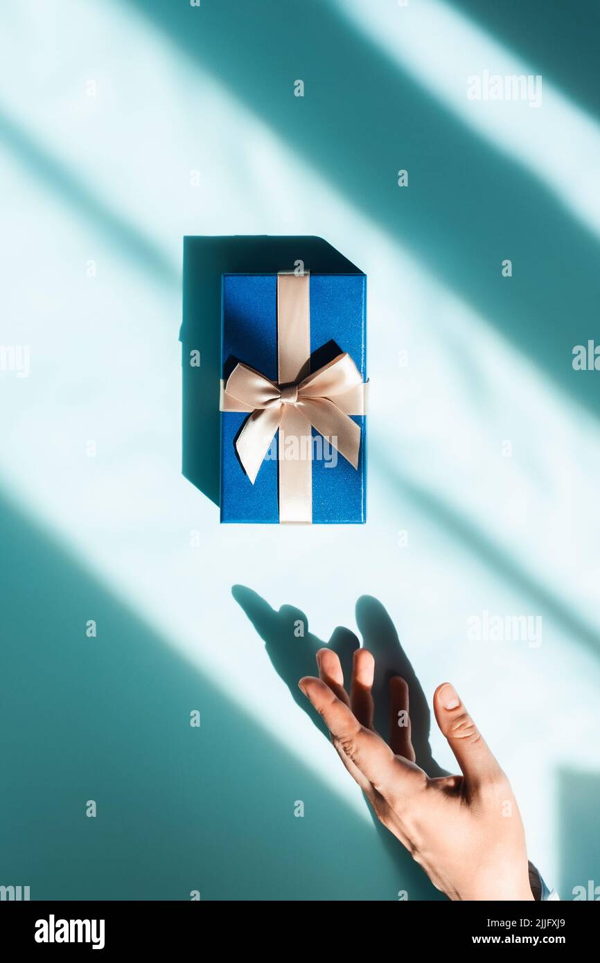 Vertical postcard with gift box in sunlight with women hand on blue background. Space for text. Stock Photo