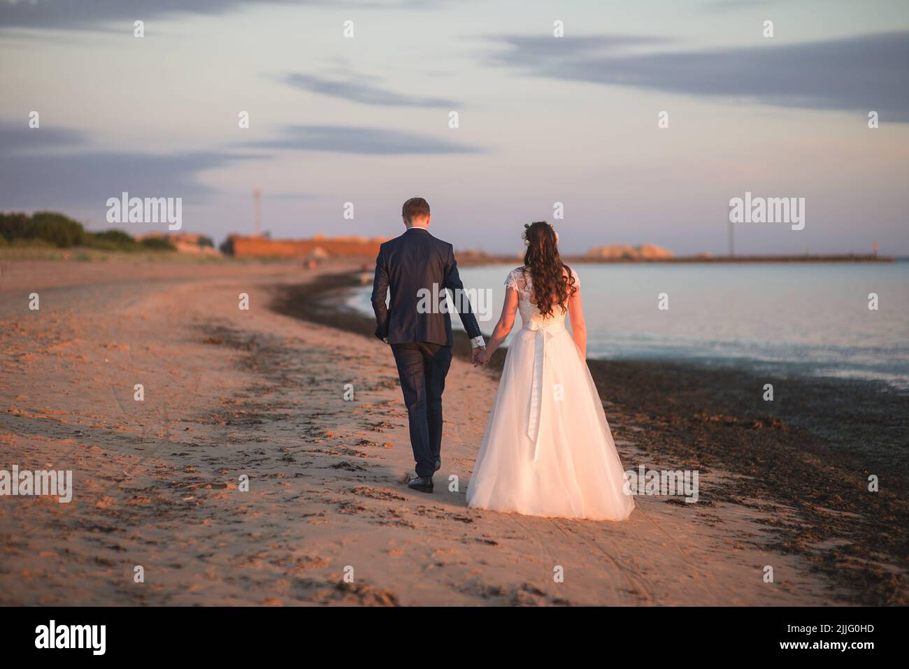 A back view of a beautiful bride and groom holding hands and walking on ...